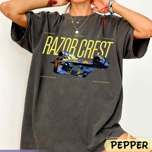May include: Dark gray t-shirt featuring the text "RAZOR CREST" in yellow, with a blue and yellow spaceship graphic. The shirt also includes additional text such as "HYPERSPACE CAPABILITIES" and "BOUNTY HUNTER TRANSPORT".