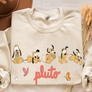 Könnte beinhalten: Cremefarbenes Sweatshirt mit einem Pluto-Cartoon-Design. Das Design zeigt Pluto in verschiedenen Posen, mit dem Wort "pluto" in Schreibschrift. Rosa und orangefarbene Schmetterlinge und eine Knochengrafik sind ebenfalls enthalten.