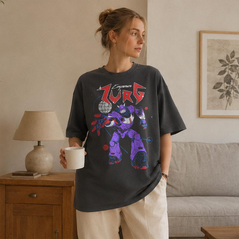 Puede incluir: Camiseta gris oscuro con un gr&aacute;fico de robot morado y el texto "Emperor Zurg". El robot tiene detalles en rojo y azul. La camiseta es de corte holgado y tiene mangas cortas.