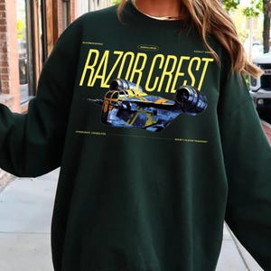 May include: Dark green sweatshirt featuring the text "RAZOR CREST" in yellow, with an illustration of a spaceship. Additional text includes "DECOMMISSIONED", "MANDALORIAN", "ASSAULT GUNSHIP", "HYPERSPACE CAPABILITIES", and "BOUNTY HUNTER TRANSPORT".
