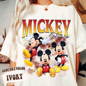 May include: Ivory Comfort Color t-shirt with a graphic of Mickey Mouse in red, yellow, and black. The shirt has the words "Mickey Mouse" in a retro font. The shirt also has the words "Comfort Color" and "Ivory" printed on the bottom left.