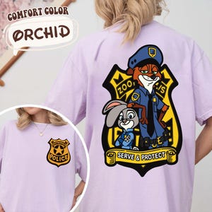 May include: Lilac t-shirt with a cartoon illustration of a fox and a rabbit in police uniforms, with the text "Zootopia Police" and "Serve & Protect". A smaller police badge is also visible on the front of the shirt.