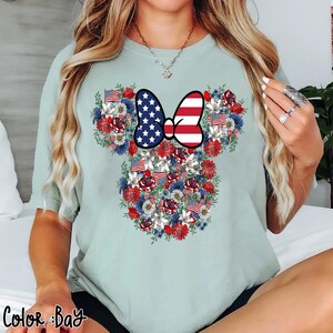 May include: A light green t-shirt featuring a floral design in the shape of a mouse head. The design incorporates red, white, and blue flowers, American flags, and a bow with a stars and stripes pattern. The text "Color Bay" is at the bottom.