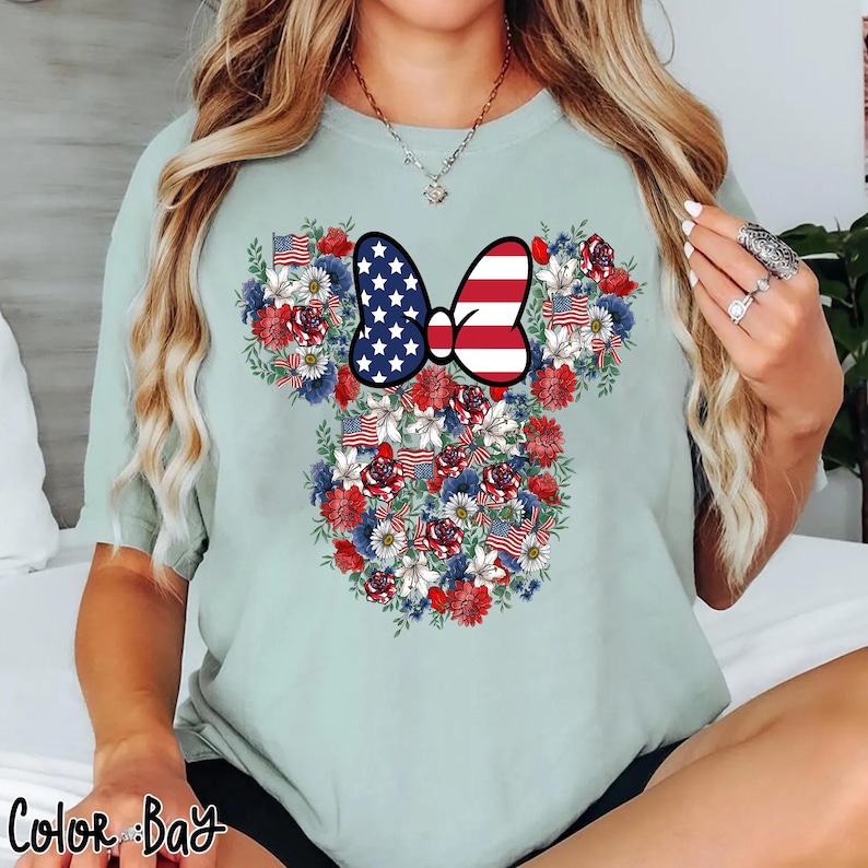 May include: A light green t-shirt featuring a floral design in the shape of a mouse head. The design incorporates red, white, and blue flowers, American flags, and a bow with a stars and stripes pattern. The text "Color Bay" is at the bottom.