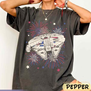 Puede incluir: Camiseta gris oscuro con una ilustración detallada de una nave espacial con detalles en rojo, blanco y azul, rodeada de fuegos artificiales y estrellas. La camiseta tiene un corte holgado y un aspecto vintage. La palabra "PEPPER" está abajo.