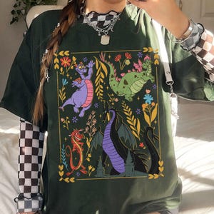 May include: Dark green t-shirt featuring a colorful graphic of cartoon dragons and floral elements. The design includes a purple dragon, a green dragon, a red dragon, and a black dragon. The shirt has short sleeves and a relaxed fit.
