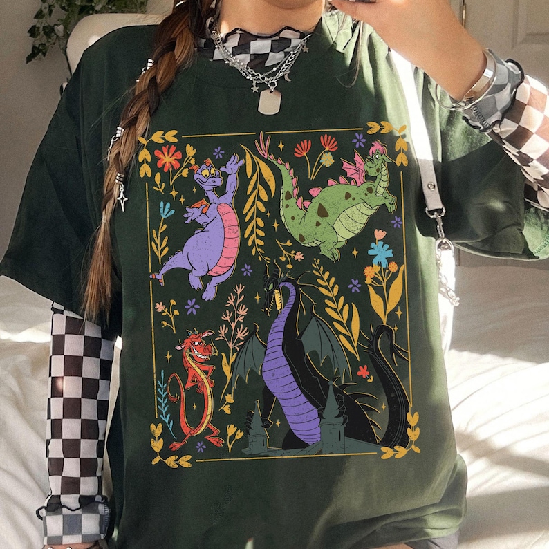 May include: Dark green t-shirt featuring a colorful graphic of cartoon dragons and floral elements. The design includes a purple dragon, a green dragon, a red dragon, and a black dragon. The shirt has short sleeves and a relaxed fit.