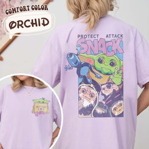 May include: Orchid-colored t-shirt with a graphic featuring a green alien character and the text "PROTECT ATTACK SNACK." The shirt also has a smaller graphic on the front. The shirt is made of Comfort Color material.