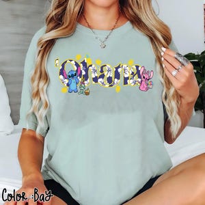Puede incluir: Una camiseta verde claro con la palabra "Ohana" en un dise&ntilde;o floral. La camiseta presenta los personajes de dibujos animados Stitch y Angel, junto con una peque&ntilde;a rana. El texto "Color Bay" est&aacute; en la parte inferior izquierda.
