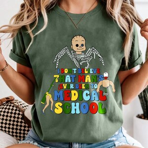 May include: Olive green t-shirt with a colorful graphic. The graphic features a cartoon spider-like figure and the text "I DON'T BELIEVE THAT MAN'S EVER BEEN TO MEDICAL SCHOOL" in various colors. The shirt has short sleeves.