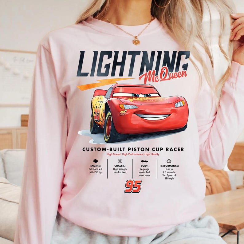 Puede incluir: Sudadera rosa claro de manga larga con un gr&aacute;fico de Lightning McQueen, un coche de carreras rojo con el n&uacute;mero 95. El texto "LIGHTNING McQueen" est&aacute; encima del coche. Texto adicional detalla las especificaciones del coche.