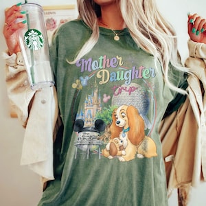 May include: An olive green t-shirt with the words "Mother Daughter Trip" in a colourful design. The graphic includes a cartoon of Lady and the Tramp, a castle, and a water tower. The shirt is a relaxed, casual style.