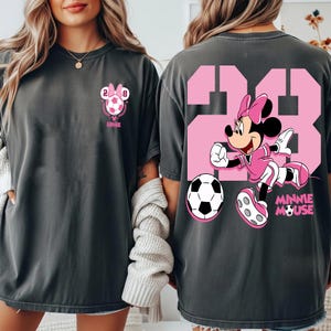 May include: Dark grey t-shirts with a Minnie Mouse soccer theme. The front shows a Minnie Mouse soccer design with the number 28. The back features a large pink number 23 and Minnie Mouse playing football.