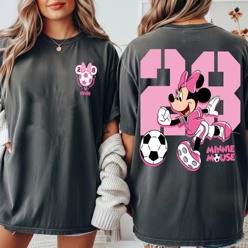 May include: Dark grey t-shirts with a Minnie Mouse soccer theme. The front shows a Minnie Mouse soccer design with the number 28. The back features a large pink number 23 and Minnie Mouse playing football.