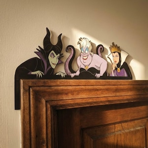 May include: Wooden cutouts of Disney villains Maleficent, Ursula, and the Evil Queen. Maleficent is in a black robe with a purple collar, Ursula has purple tentacles, and the Evil Queen wears a crown. The cutouts are placed above a wooden surface.