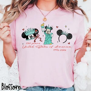 May include: Pink t-shirt featuring a graphic design celebrating the United States of America's 250th anniversary. The design includes Mickey Mouse as the Statue of Liberty, a Mickey Mouse ear headband, and the text "250 years United States of America 1776-2026."
