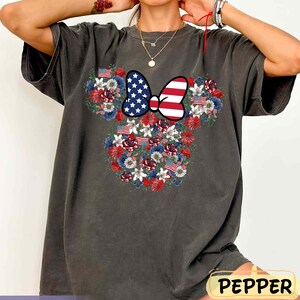 May include: Dark gray t-shirt featuring a floral design in the shape of a mouse head. The design includes a bow with the American flag and flowers in red, white, and blue. The word "PEPPER" is in a yellow rectangle.