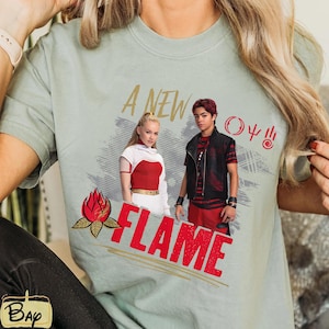 May include: A pale green t-shirt with a design featuring two people and the words "A NEW FLAME" in gold and red. The design includes a red flame graphic and other symbols. A casual wear item.
