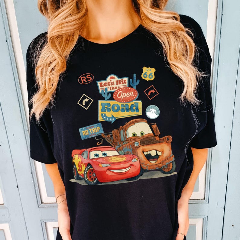 May include: Black t-shirt with a graphic of Lightning McQueen and Mater from the film Cars. The design includes the text "Let's Hit the Open Road" and Route 66 imagery. The shirt has a crew neck and short sleeves.