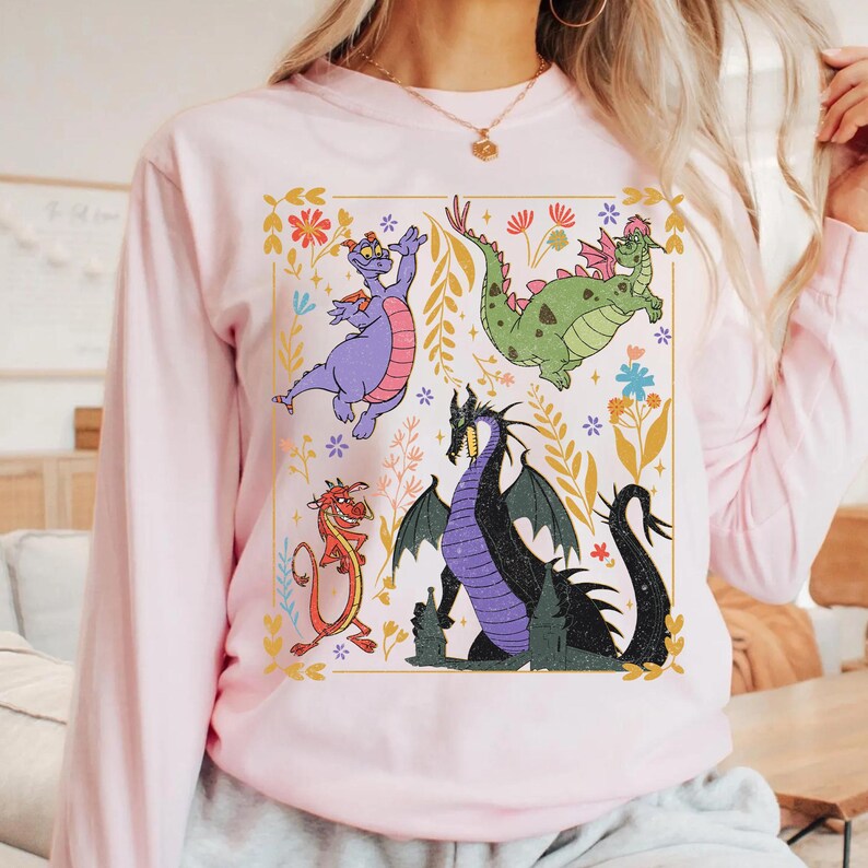 May include: A light pink long-sleeve sweatshirt featuring a colorful graphic of four cartoon dragons within a gold-lined square. The dragons are purple, green, red, and black, surrounded by floral and leaf designs.