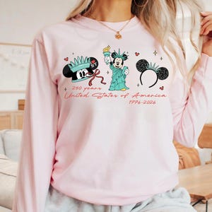 May include: A light pink long-sleeved sweatshirt featuring three Mickey Mouse-themed designs. The designs include Mickey Mouse with a crown, as the Statue of Liberty, and a pair of mouse ears. Text reads "250 years United States of America 1776-2026."