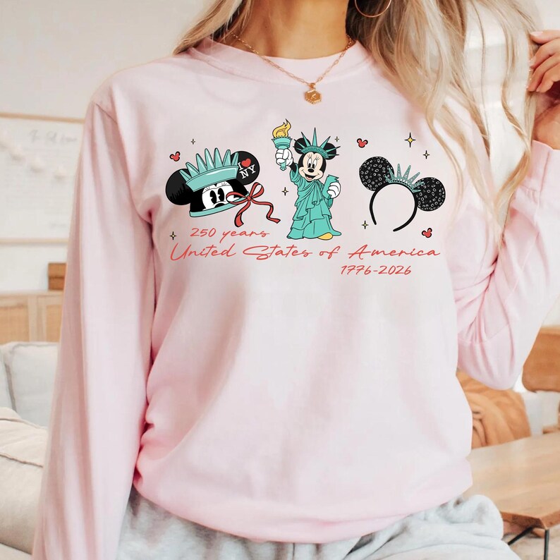 May include: A light pink long-sleeved sweatshirt featuring three Mickey Mouse-themed designs. The designs include Mickey Mouse with a crown, as the Statue of Liberty, and a pair of mouse ears. Text reads "250 years United States of America 1776-2026."