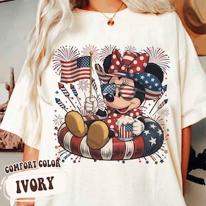 May include: Ivory Comfort Color t-shirt featuring a cartoon character in patriotic attire, holding an American flag and a mug. The character is wearing sunglasses and a bow, surrounded by fireworks and sitting in a red, white, and blue inner tube.
