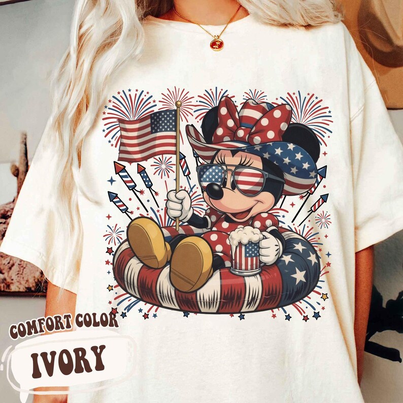May include: Ivory Comfort Color t-shirt featuring a cartoon character in patriotic attire, holding an American flag and a mug. The character is wearing sunglasses and a bow, surrounded by fireworks and sitting in a red, white, and blue inner tube.