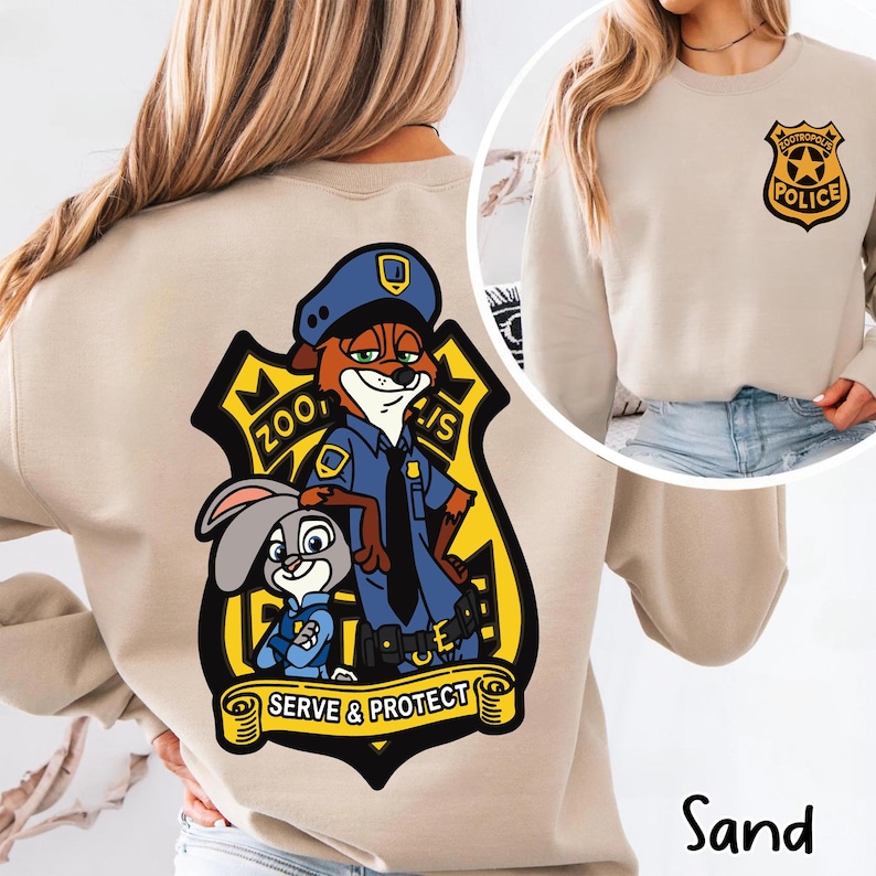 May include: Sand-coloured sweatshirt with a Zootropolis police badge design. The graphic features Nick Wilde and Judy Hopps, with the text "Zootropolis" and "Serve & Protect". A smaller badge is on the front. The word "Sand" is at the bottom.