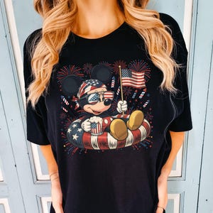 May include: Black t-shirt featuring a cartoon Mickey Mouse design celebrating Independence Day. Mickey is wearing sunglasses and a patriotic bandana, holding a mug and a small American flag while floating in a red, white, and blue inner tube. Fireworks are in the background.