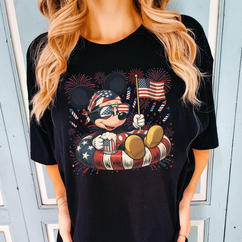 May include: Black t-shirt featuring a cartoon Mickey Mouse design celebrating Independence Day. Mickey is wearing sunglasses and a patriotic bandana, holding a mug and a small American flag while floating in a red, white, and blue inner tube. Fireworks are in the background.