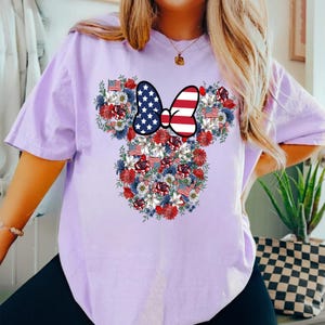 May include: A light purple t-shirt featuring a floral design in the shape of a mouse head. The design incorporates red, white, and blue flowers, American flags, and a bow with a stars and stripes pattern.