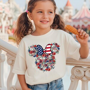 May include: A cream-colored t-shirt featuring a floral Minnie Mouse design with red, white, and blue accents. The design includes a bow with a stars and stripes pattern and small American flags. The shirt is worn by a young girl.