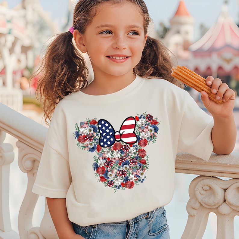 May include: A cream-colored t-shirt featuring a floral Minnie Mouse design with red, white, and blue accents. The design includes a bow with a stars and stripes pattern and small American flags. The shirt is worn by a young girl.
