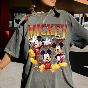 May include: Grey t-shirt with a graphic of Mickey Mouse in three poses. The text "MICKEY Mouse" is printed in orange and yellow. The cartoon characters are wearing red shorts and yellow shoes. The t-shirt is a casual style.