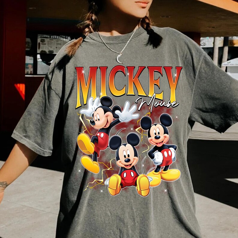 May include: Grey t-shirt with a graphic of Mickey Mouse in three poses. The text "MICKEY Mouse" is printed in orange and yellow. The cartoon characters are wearing red shorts and yellow shoes. The t-shirt is a casual style.