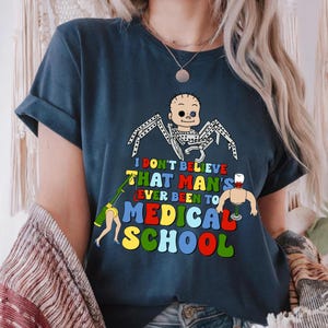 May include: Navy blue t-shirt with a colorful graphic. The graphic features a baby-like figure with spider legs, a detached arm, and the text "I DON'T BELIEVE THAT MAN'S EVER BEEN TO MEDICAL SCHOOL."