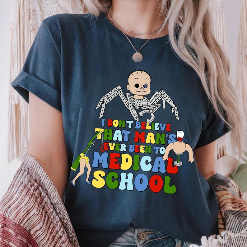 May include: Navy blue t-shirt with a colorful graphic. The graphic features a baby-like figure with spider legs, a detached arm, and the text "I DON'T BELIEVE THAT MAN'S EVER BEEN TO MEDICAL SCHOOL."