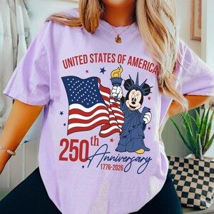 May include: A light purple t-shirt with a graphic celebrating the 250th anniversary of the United States. The design includes the American flag, a cartoon character dressed as the Statue of Liberty, and the text "United States of America".