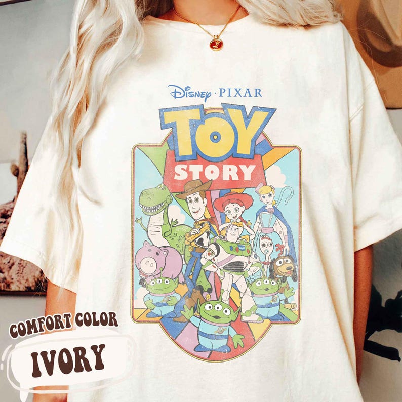 Puede incluir: Camiseta de color marfil con un gr&aacute;fico colorido de personajes de la pel&iacute;cula de Disney Pixar, Toy Story. La camiseta tiene las palabras "Comfort Color" e "Ivory" impresas en la parte inferior izquierda.