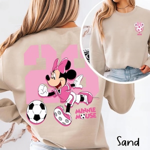 May include: Sand-coloured sweatshirt with a large pink number 24 and a Minnie Mouse graphic in a soccer uniform. The design includes a soccer ball and the words "Minnie Mouse". A smaller image of Minnie with a soccer ball is on the upper right.