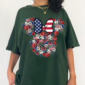 May include: Dark green t-shirt featuring a floral Minnie Mouse design. The design incorporates red, white, and blue flowers, American flags, and a bow with a stars and stripes pattern. The shirt is a comfortable, casual style.