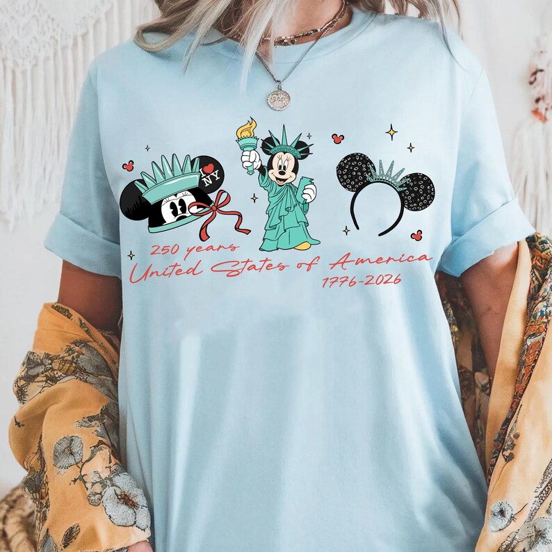 May include: Light blue t-shirt featuring Mickey Mouse illustrations in the style of the Statue of Liberty and other patriotic designs. The text reads "250 years United States of America 1776-2026". The shirt has rolled-up sleeves.