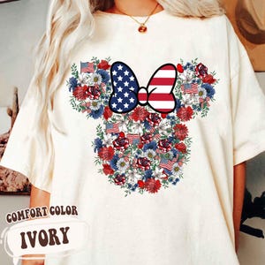 May include: Ivory-colored t-shirt featuring a floral design in the shape of a mouse head. The design incorporates red, white, and blue flowers, American flags, and a bow with a stars and stripes pattern. The text "COMFORT COLOR IVORY" is at the bottom.