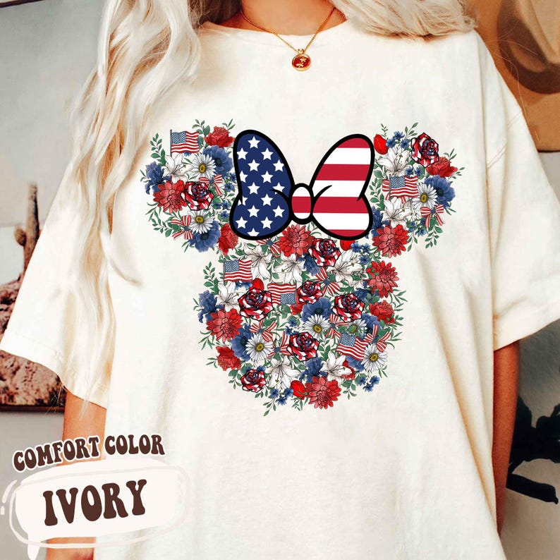 May include: Ivory-colored t-shirt featuring a floral design in the shape of a mouse head. The design incorporates red, white, and blue flowers, American flags, and a bow with a stars and stripes pattern. The text "COMFORT COLOR IVORY" is at the bottom.