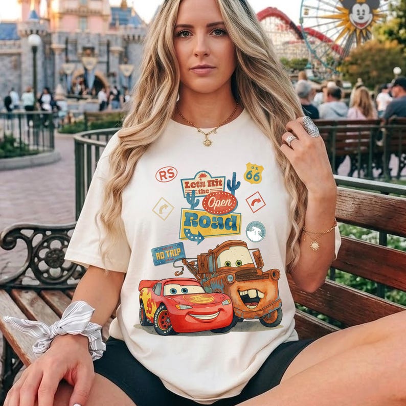 May include: Cream-coloured t-shirt featuring Lightning McQueen and Mater from the film Cars, with the text "Let's Hit the Open Road." The design includes road signs and Route 66 imagery. A fun, casual tee.