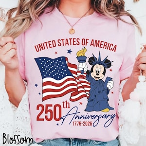 May include: Pink t-shirt with the text "UNITED STATES OF AMERICA" and "250th Anniversary 1776-2026". Features a waving American flag and a cartoon character dressed as the Statue of Liberty.