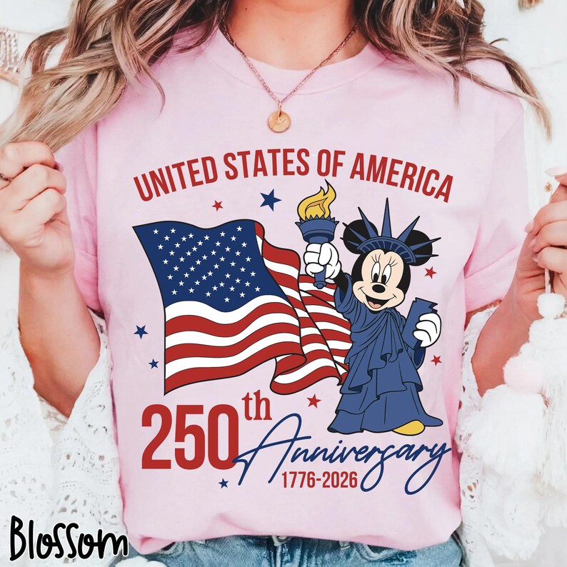 May include: Pink t-shirt with the text "UNITED STATES OF AMERICA" and "250th Anniversary 1776-2026". Features a waving American flag and a cartoon character dressed as the Statue of Liberty.