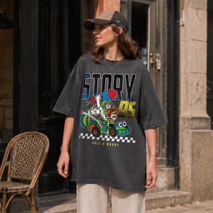 May include: Dark gray t-shirt featuring a graphic of Buzz Lightyear and Woody from Toy Story in a race car. The shirt has the word "STORY" above the graphic and "95" below it. The words "BUZZ & WOODY" are at the bottom.