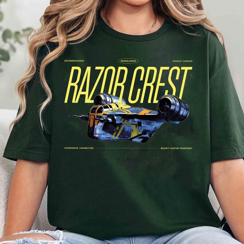 May include: Dark green t-shirt featuring a yellow and blue illustration of the Razor Crest starship from The Mandalorian. The text "RAZOR CREST" is in large yellow letters above the ship, with additional text below.